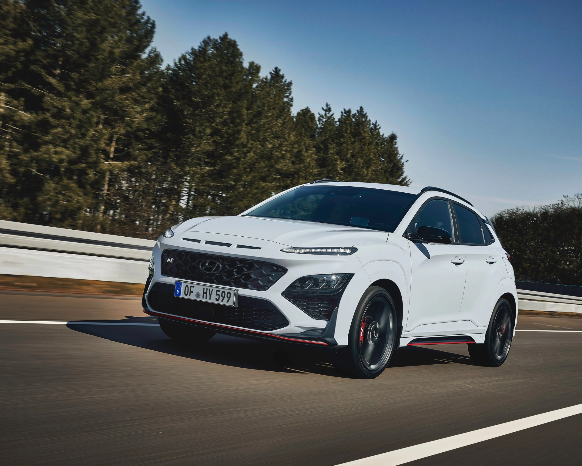 White Hyundai Kona N compact SUV with black roof and red accents driving on a road with trees in the background.