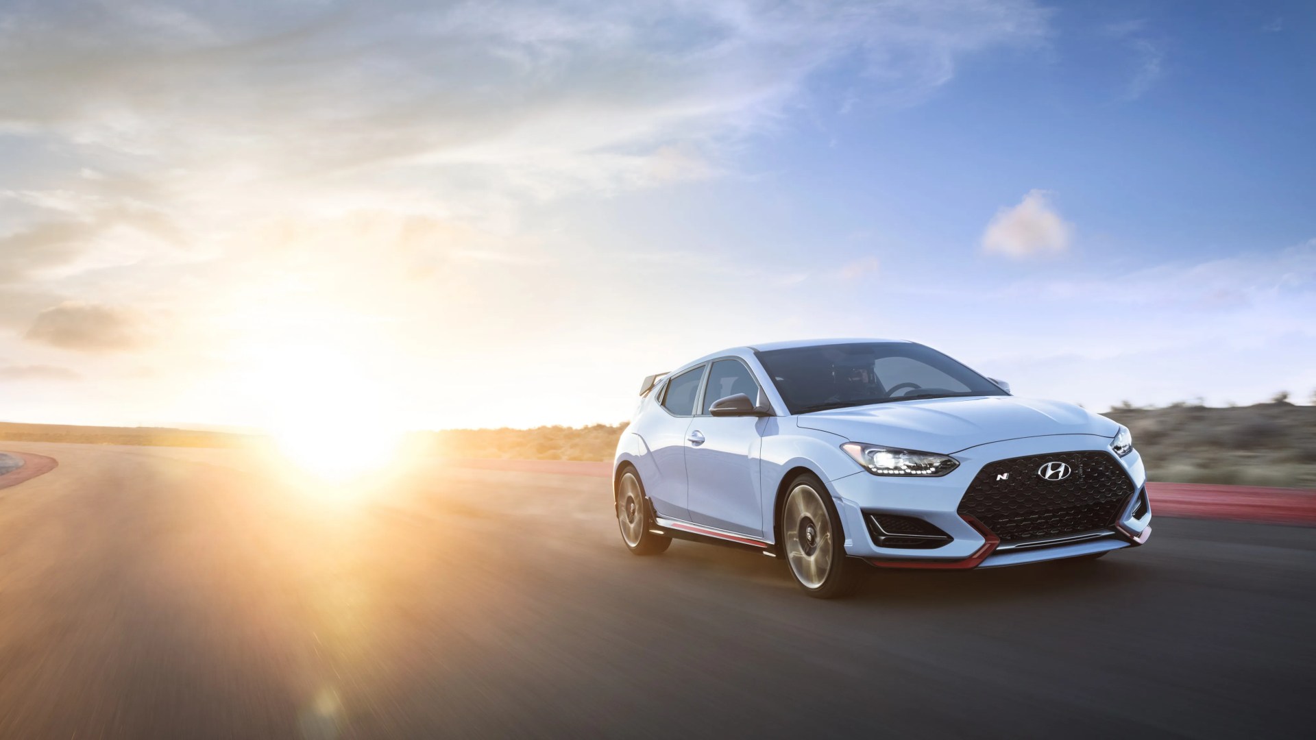 Light blue Hyundai hatchback with black grille and red accents driving on a racetrack at sunset.