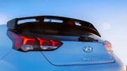 Rear view of a light blue Hyundai Veloster N with illuminated taillights and a black rear spoiler against a clear sky.