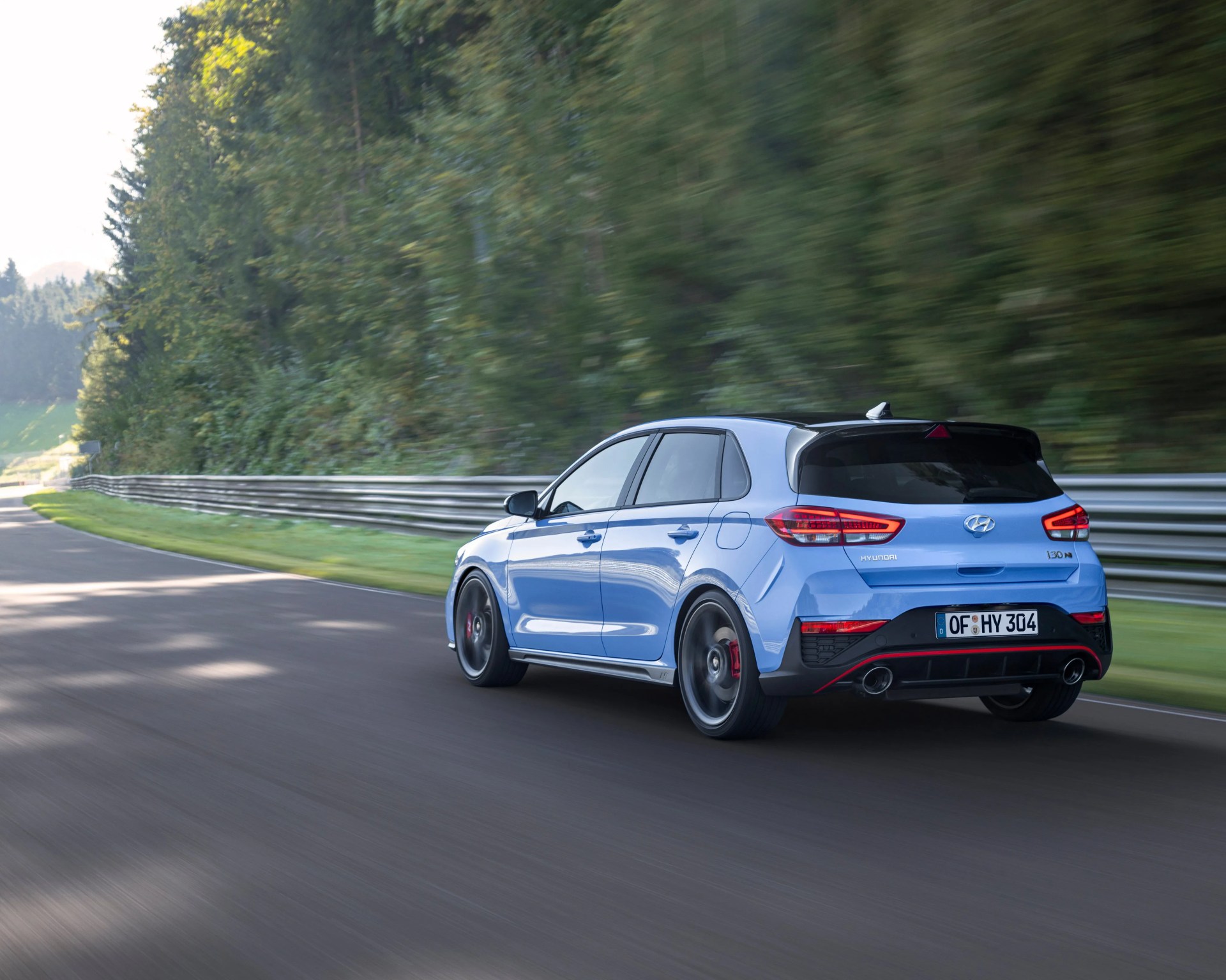 Blue Hyundai i30 N hatchback driving on a tree-lined racetrack with motion blur.