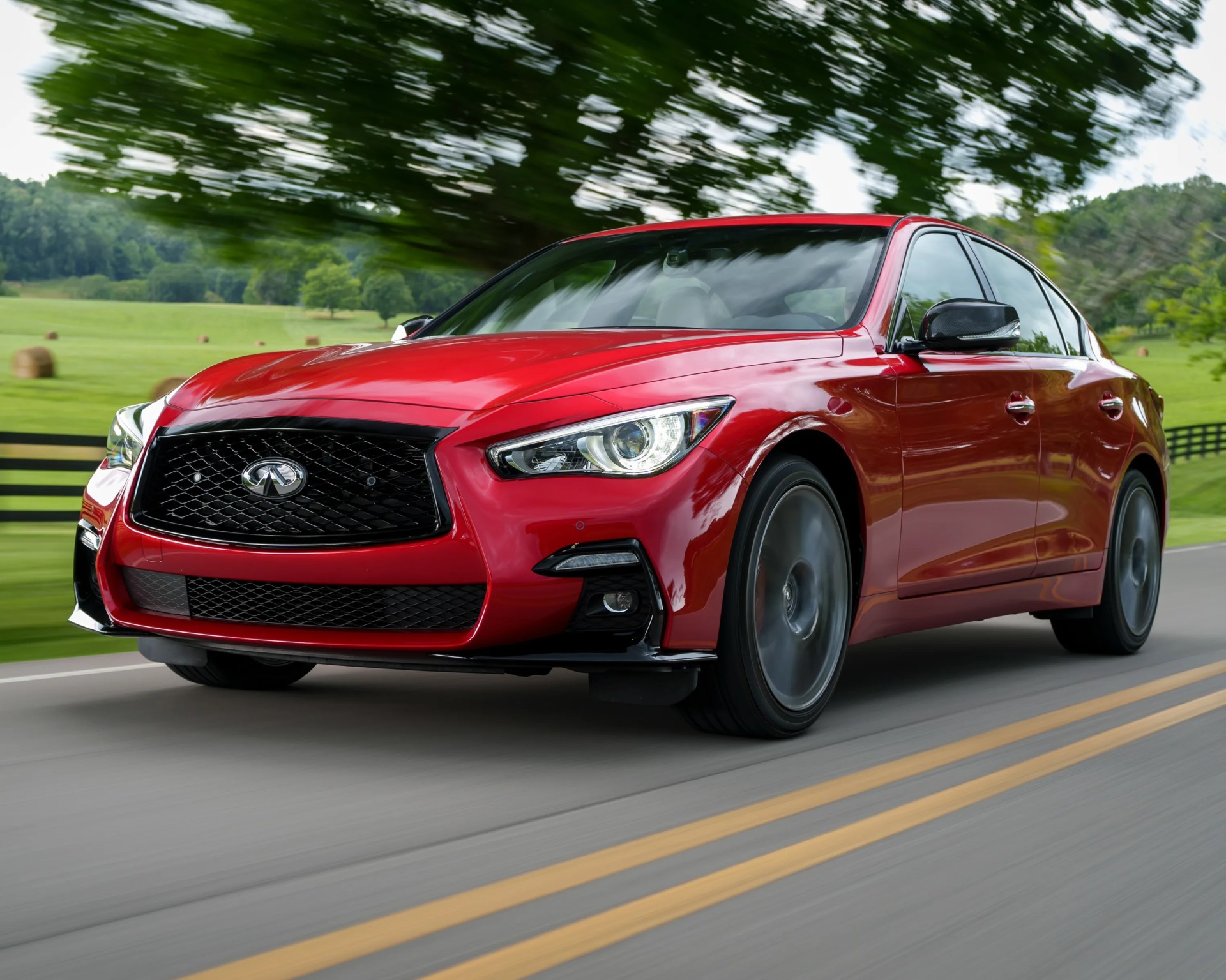 Red Infiniti sedan driving on a rural road with green fields and trees in the background.