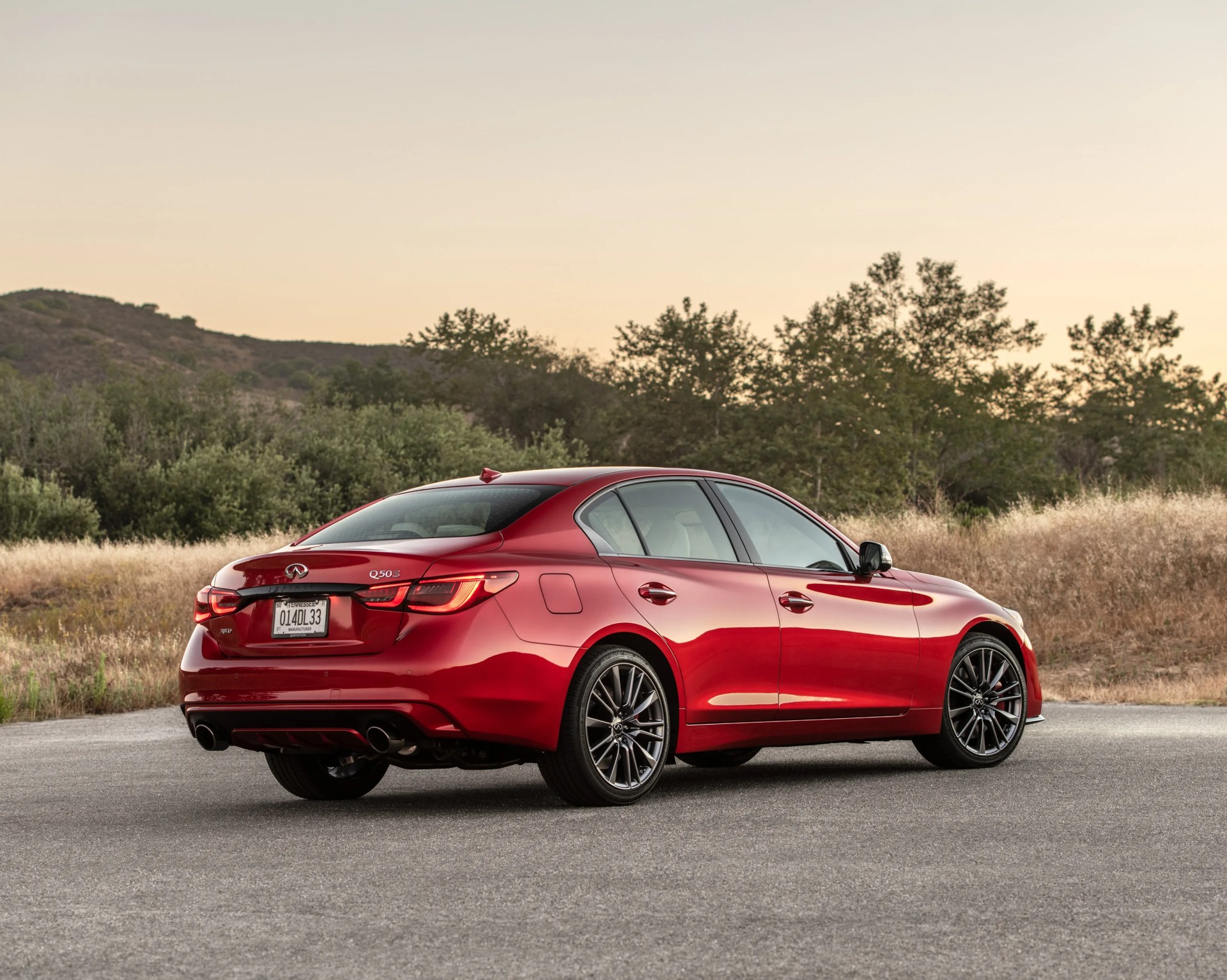 Red Infiniti Q50S sedan parked on a paved road with hills and trees in the background at sunset.