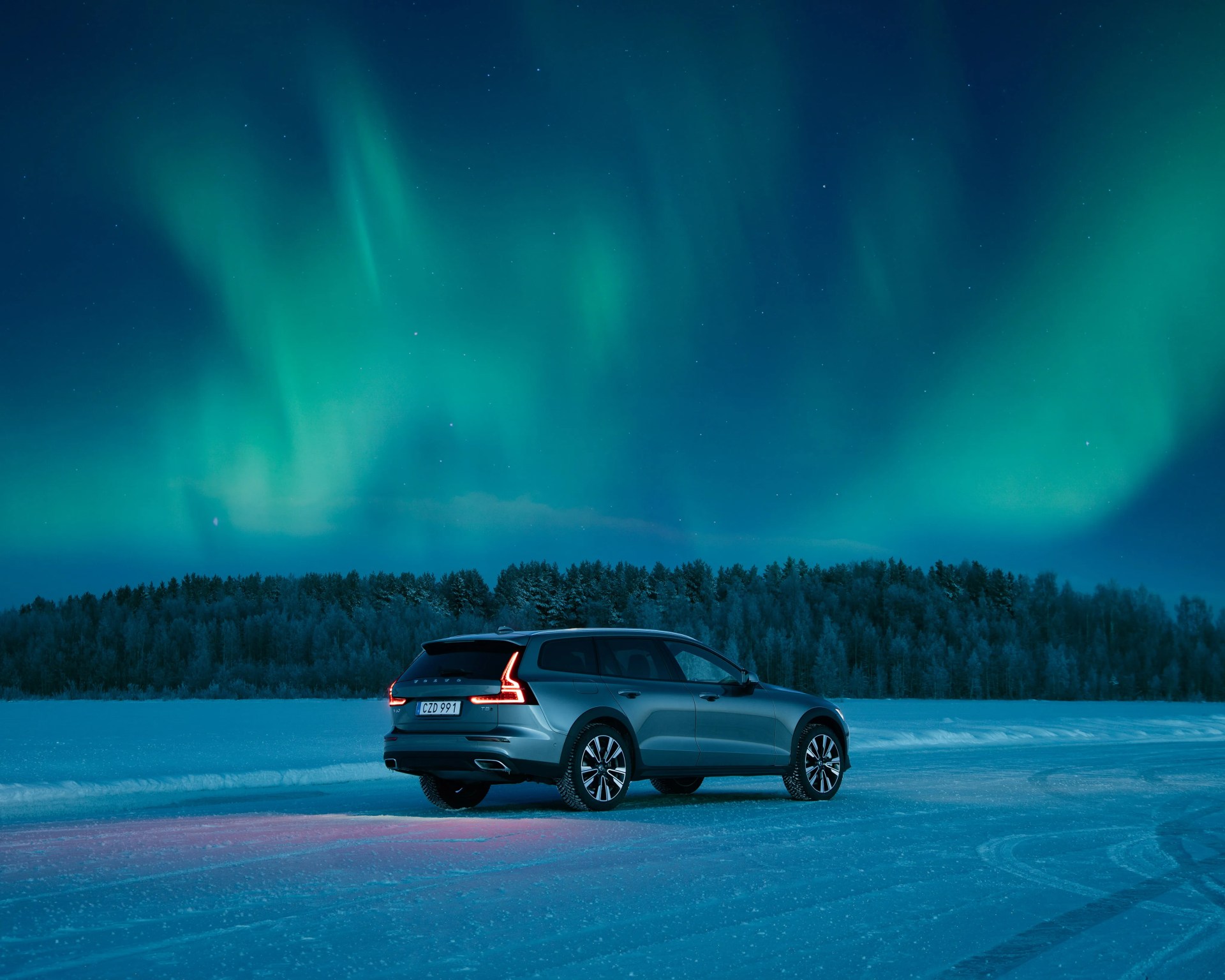 Silver Volvo V90 Cross Country station wagon on snowy ground under green northern lights at night.