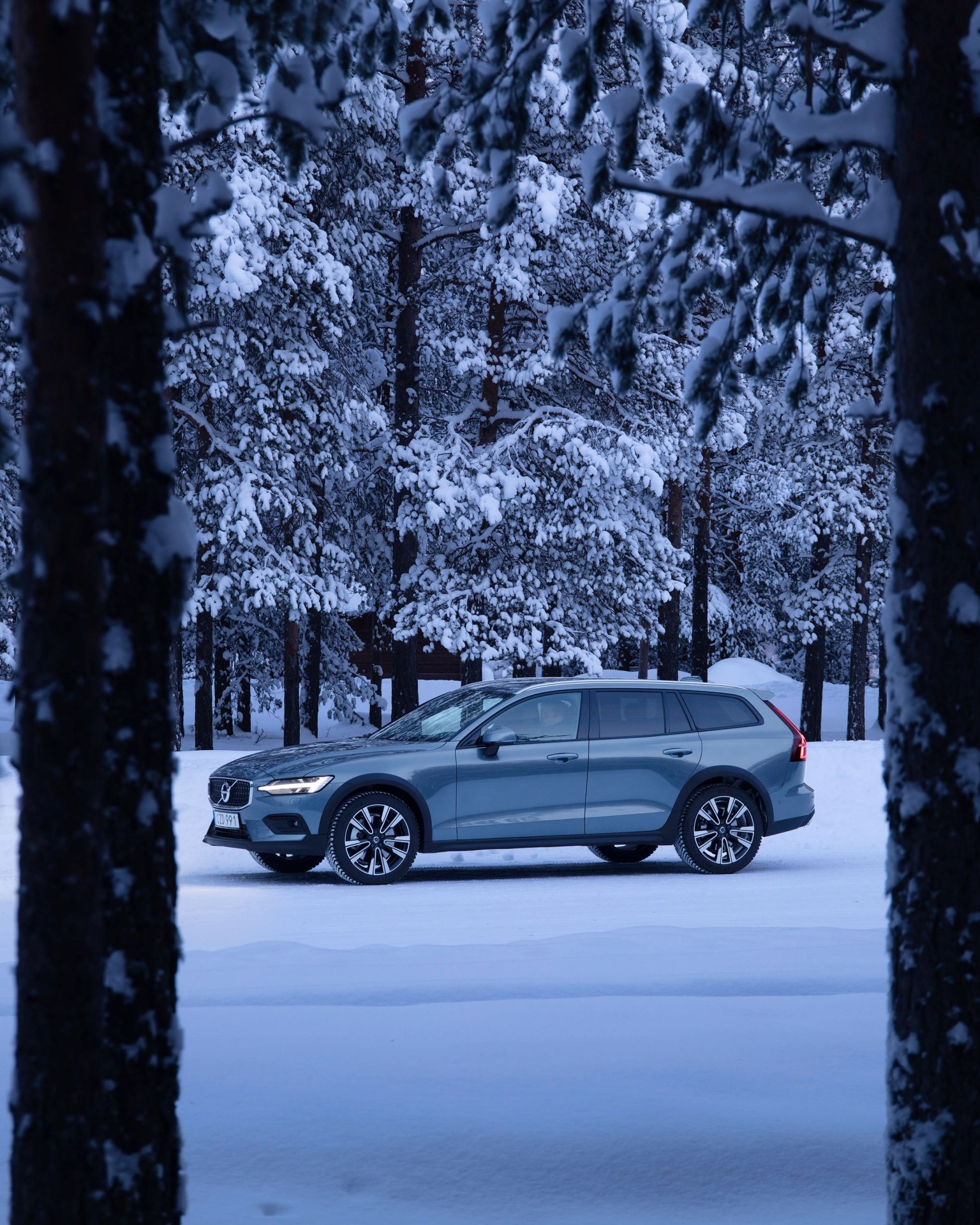 Blue Volvo station wagon parked on snow-covered ground with snow-laden pine trees in the background.