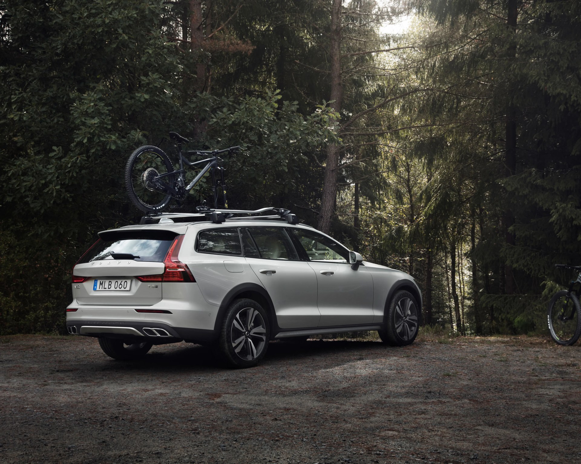 White Volvo V60 Cross Country station wagon with a mountain bike mounted on the roof rack, parked on a forest gravel clearing.