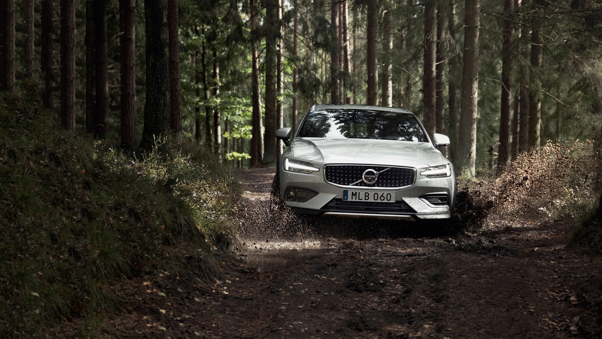Silver Volvo car splashing mud while driving on a forest dirt road.