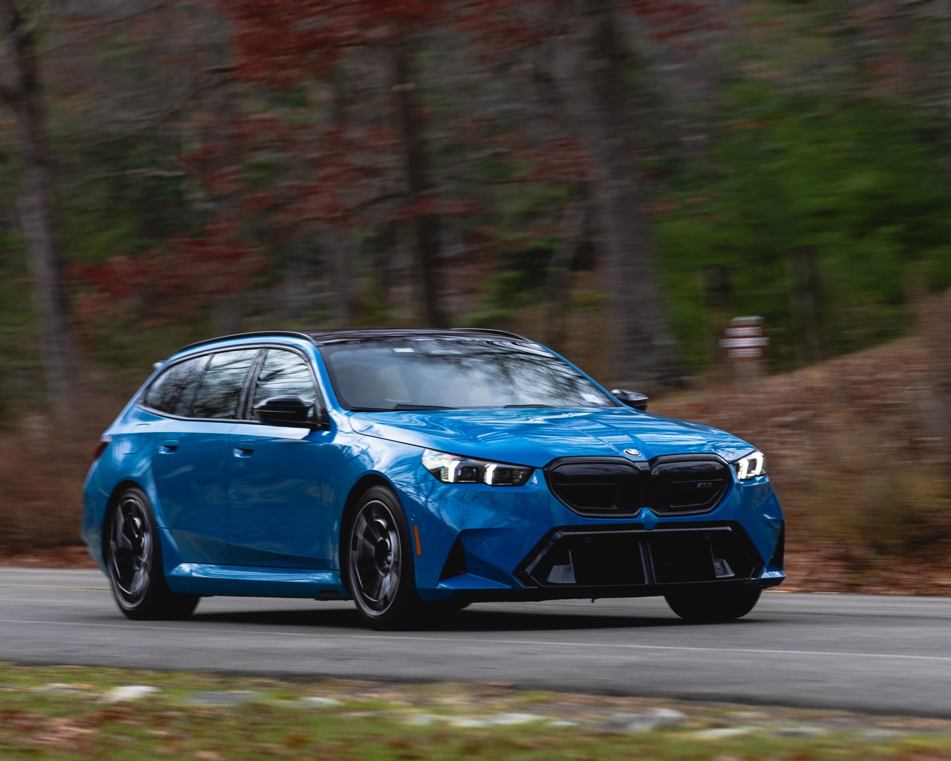 Blue BMW M3 Touring with black grille and roof driving on a forest road.