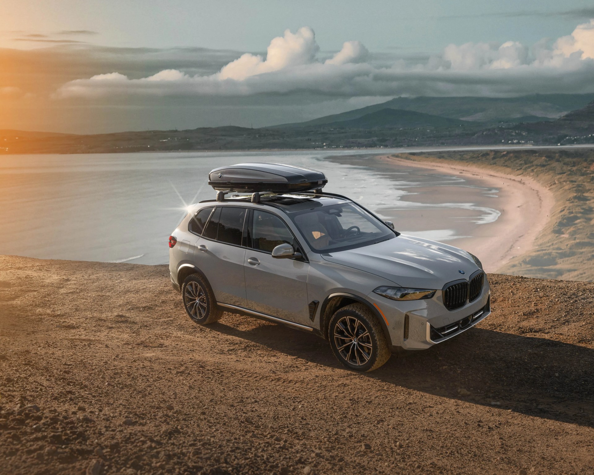 Silver BMW SUV with roof cargo box parked on a dirt cliff overlooking a beach and ocean at sunset.