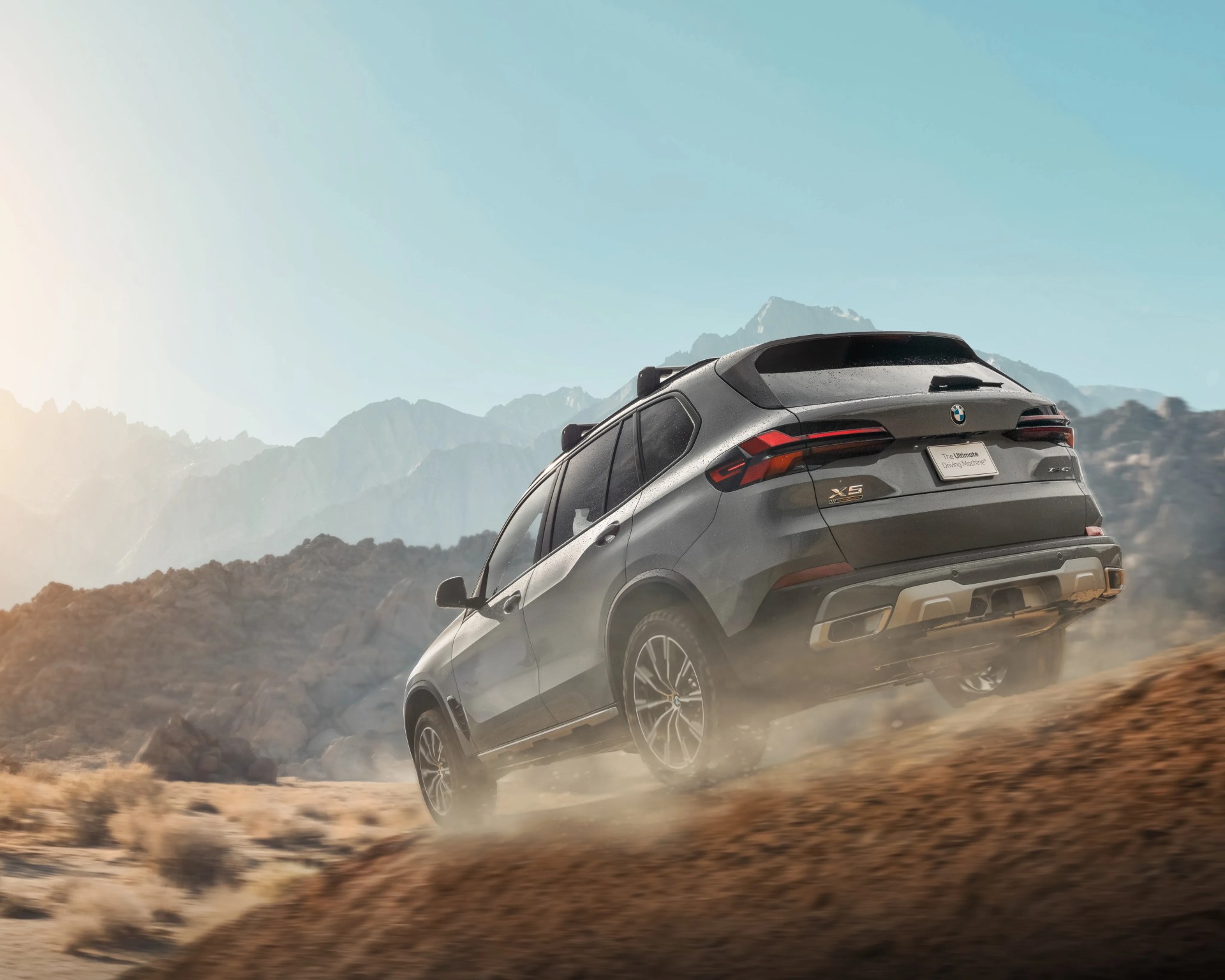 Gray BMW X5 SUV driving uphill on a dusty mountain trail with rocky terrain and clear sky.