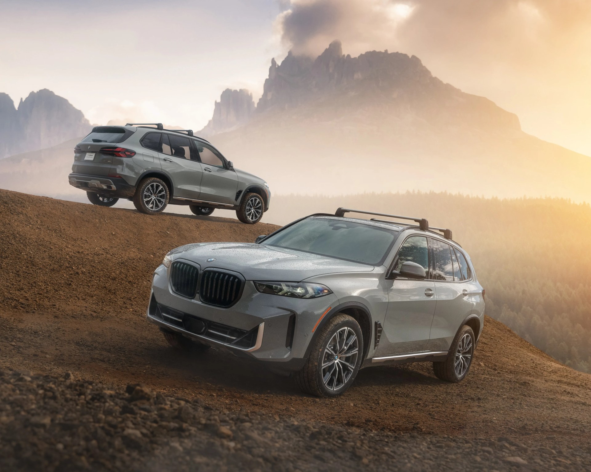 Two gray BMW SUVs on a dirt hill with mountainous forest landscape in the background at sunset.