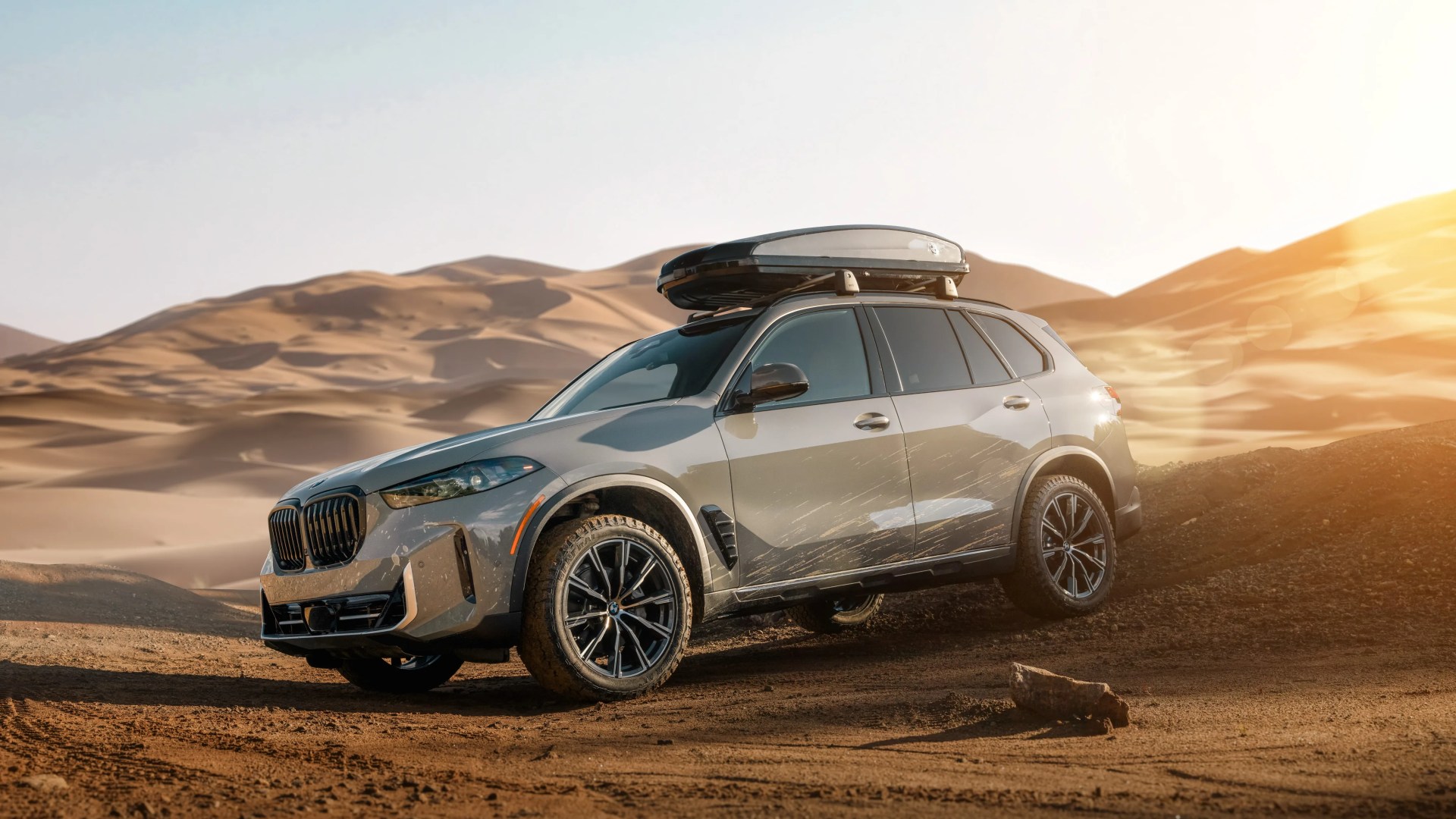 Silver BMW SUV with roof cargo box parked on dirt with sand dunes in the background at sunset.