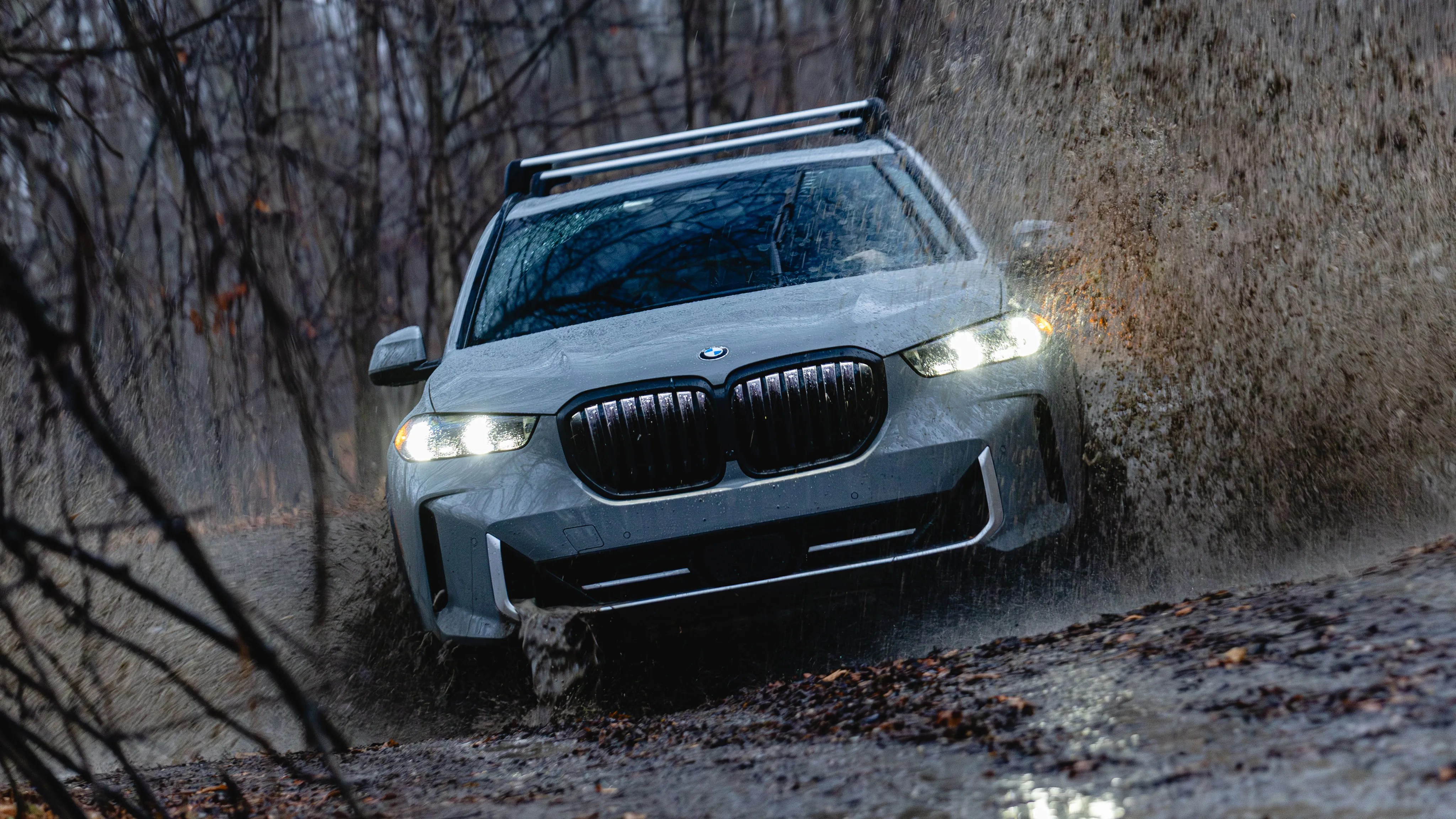 White BMW SUV splashing mud while driving on a wet forest trail with headlights on.