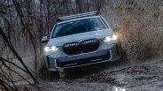 White BMW SUV splashing mud while driving on a wet forest trail with headlights on.