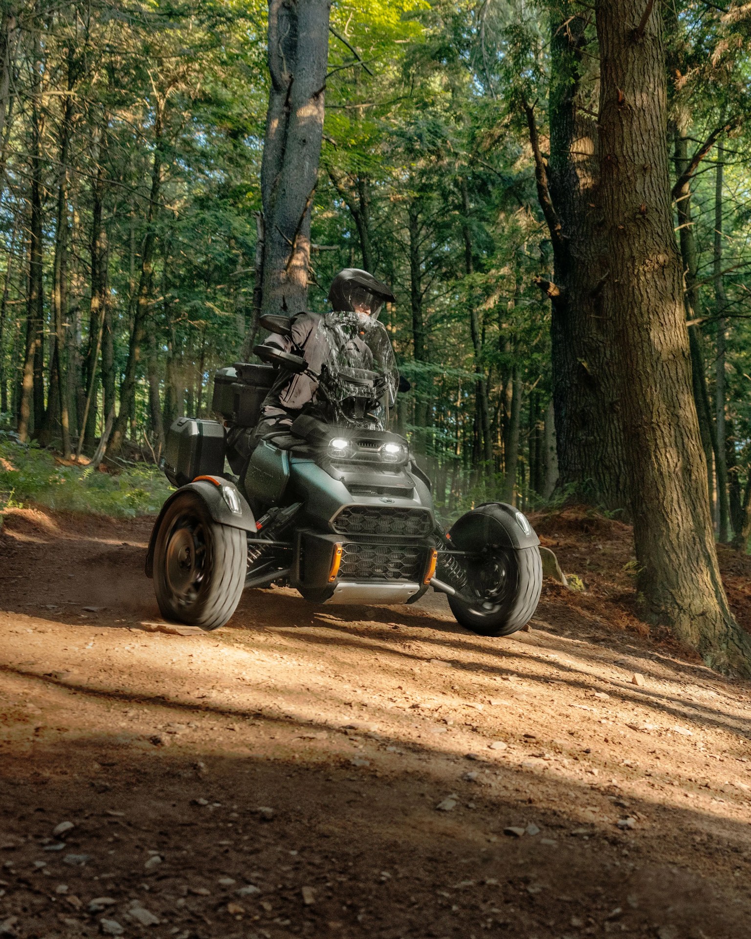 Person riding a black three-wheeled off-road motorcycle on a dirt trail in a forest.