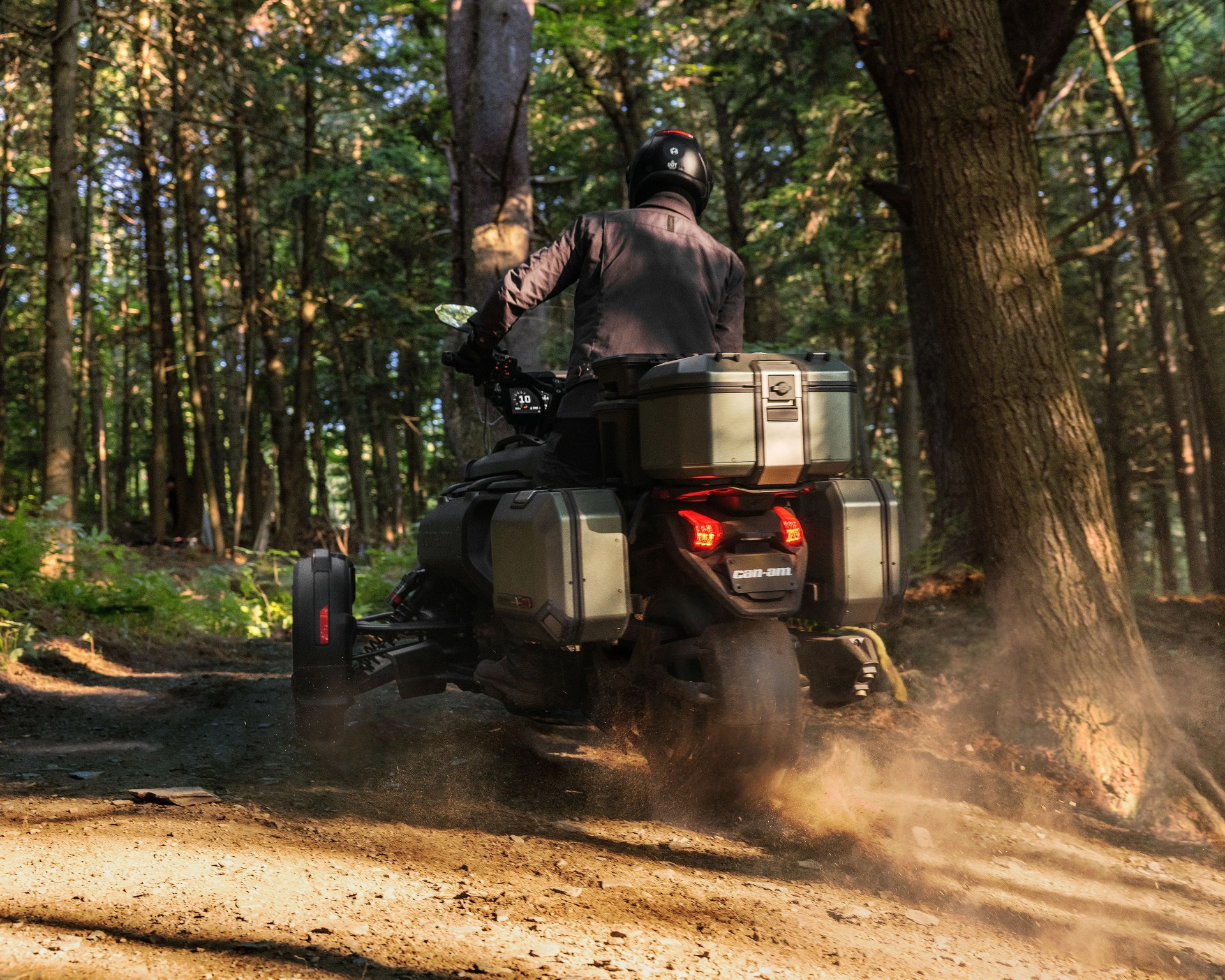 Person riding a Can-Am three-wheeled motorcycle with storage boxes on a forest trail kicking up dust.