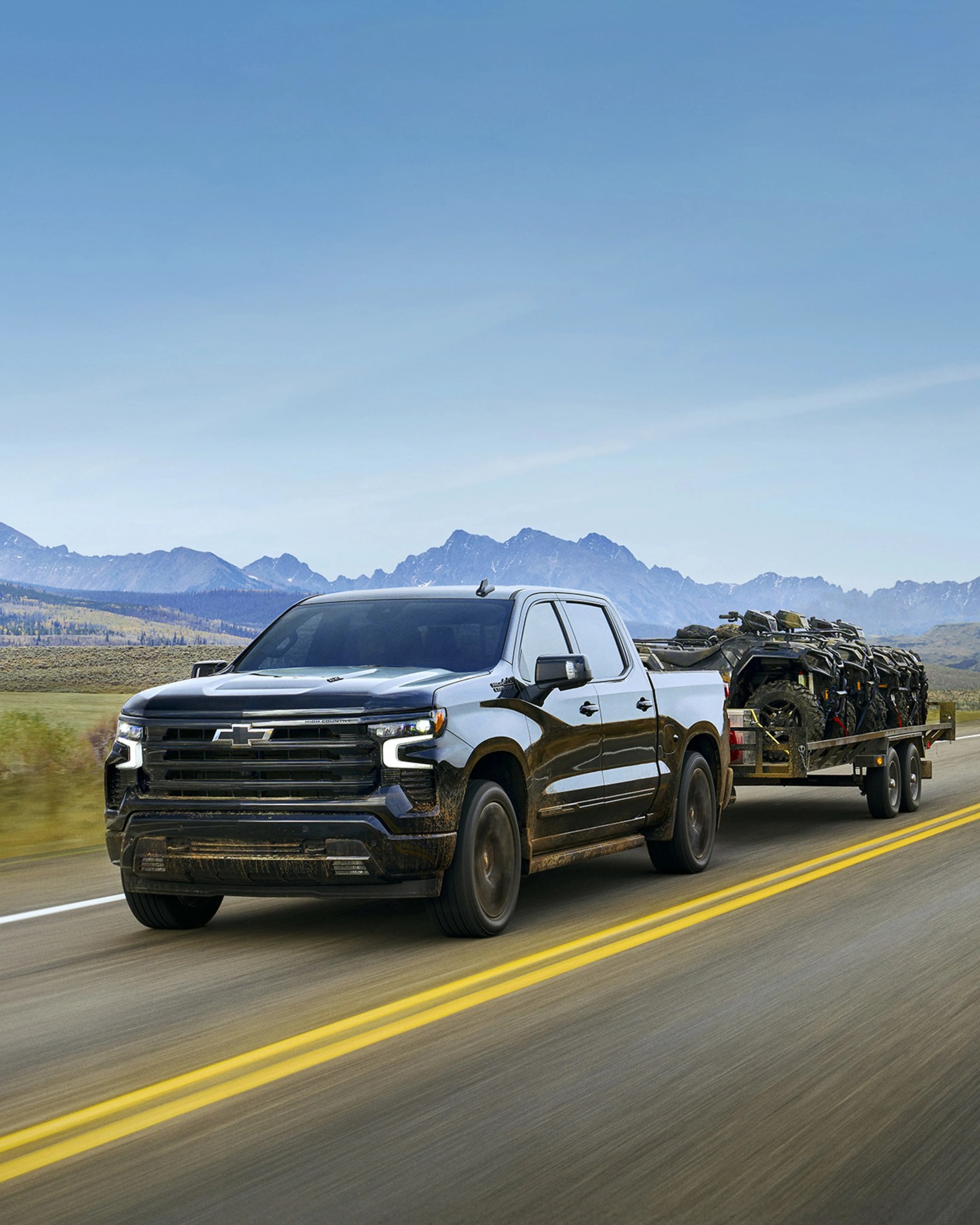 Black Chevrolet pickup truck towing a trailer loaded with ATVs on a highway with mountains in the background.