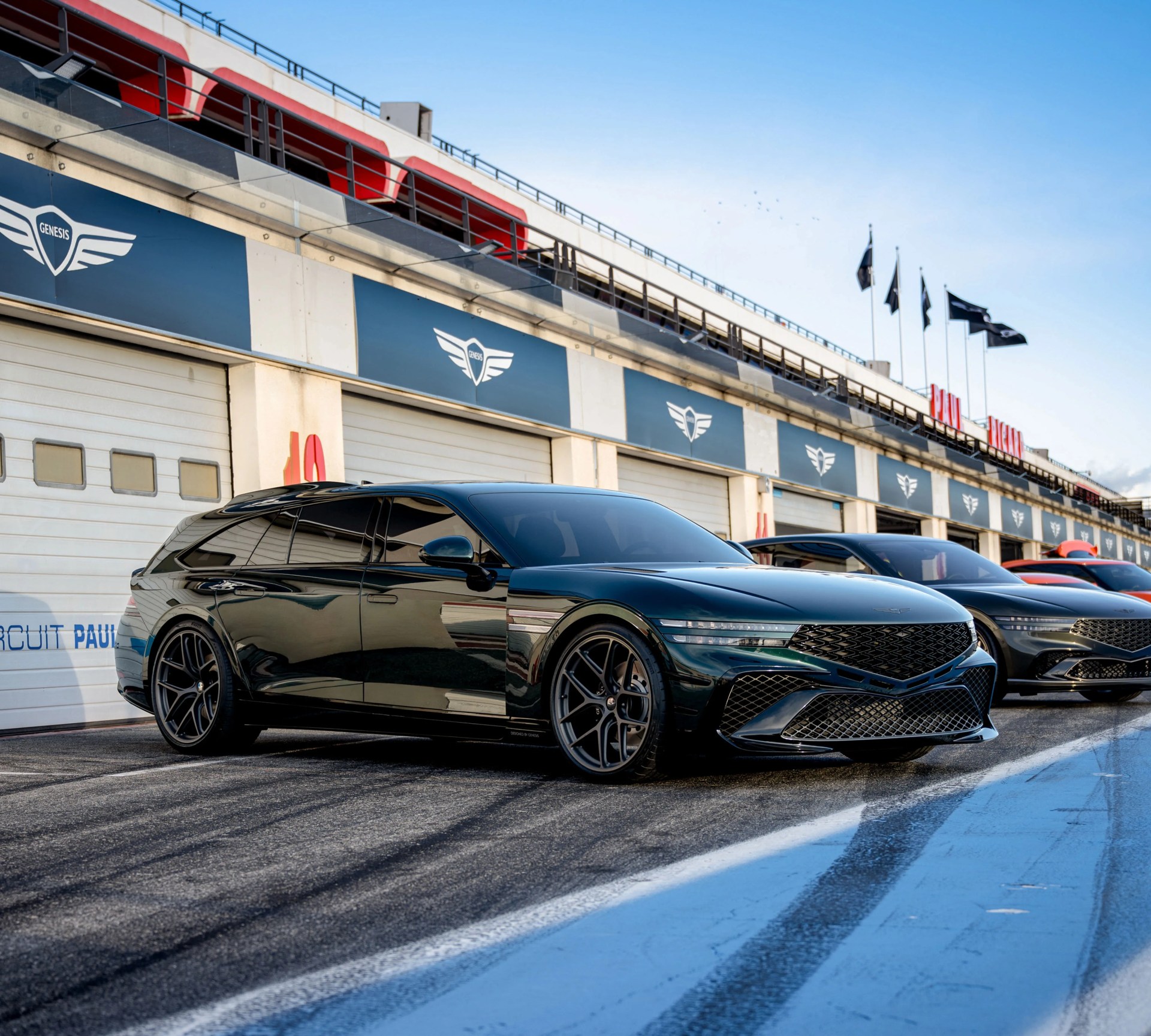 Glossy black Genesis station wagon with dark tinted windows parked on a race track pit lane.