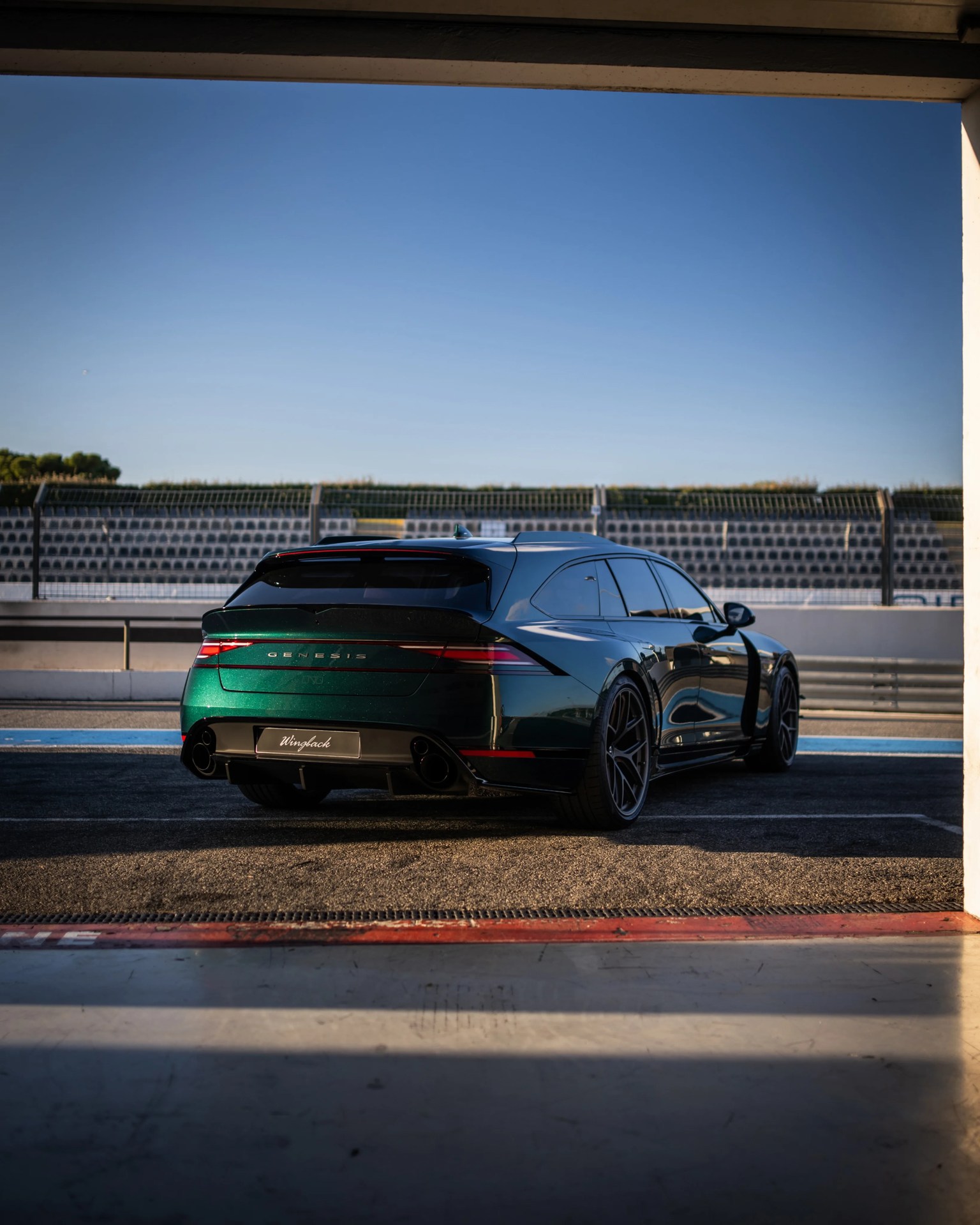Dark green Genesis station wagon with tinted windows parked on a race track under a clear blue sky.