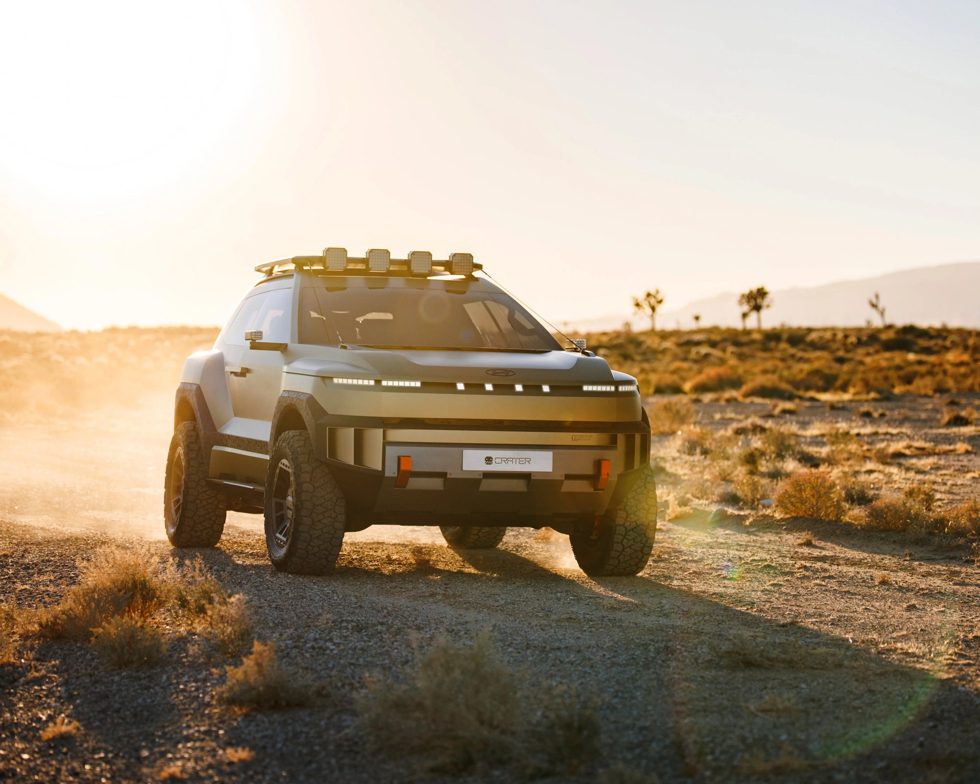 Matte gray off-road SUV with roof lights and rugged tires driving on a dusty desert trail at sunset.