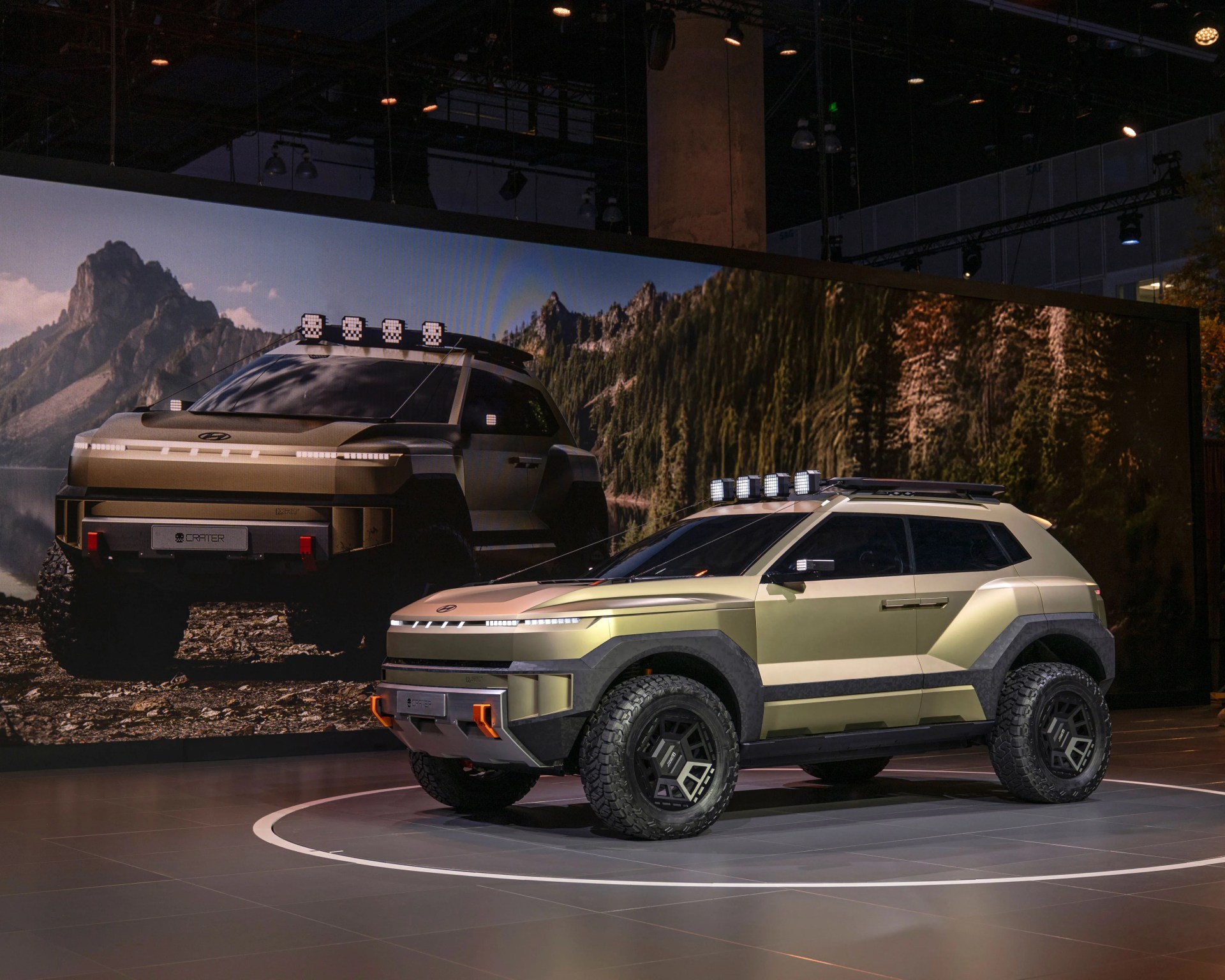 Matte gold and black Hyundai electric off-road SUV with roof lights displayed indoors in front of a large nature-themed backdrop.