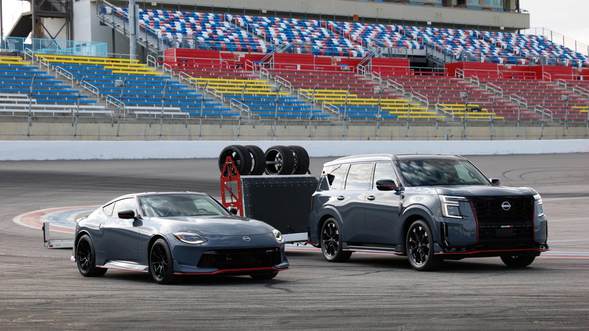 Two dark gray Nissan Nismo vehicles, a sports car and an SUV, parked on a racetrack with a tire rack trailer between them.