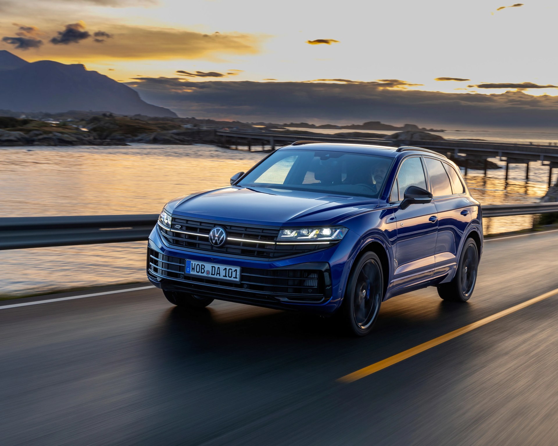Blue Volkswagen SUV driving on a coastal road at sunset with mountains and water in the background.