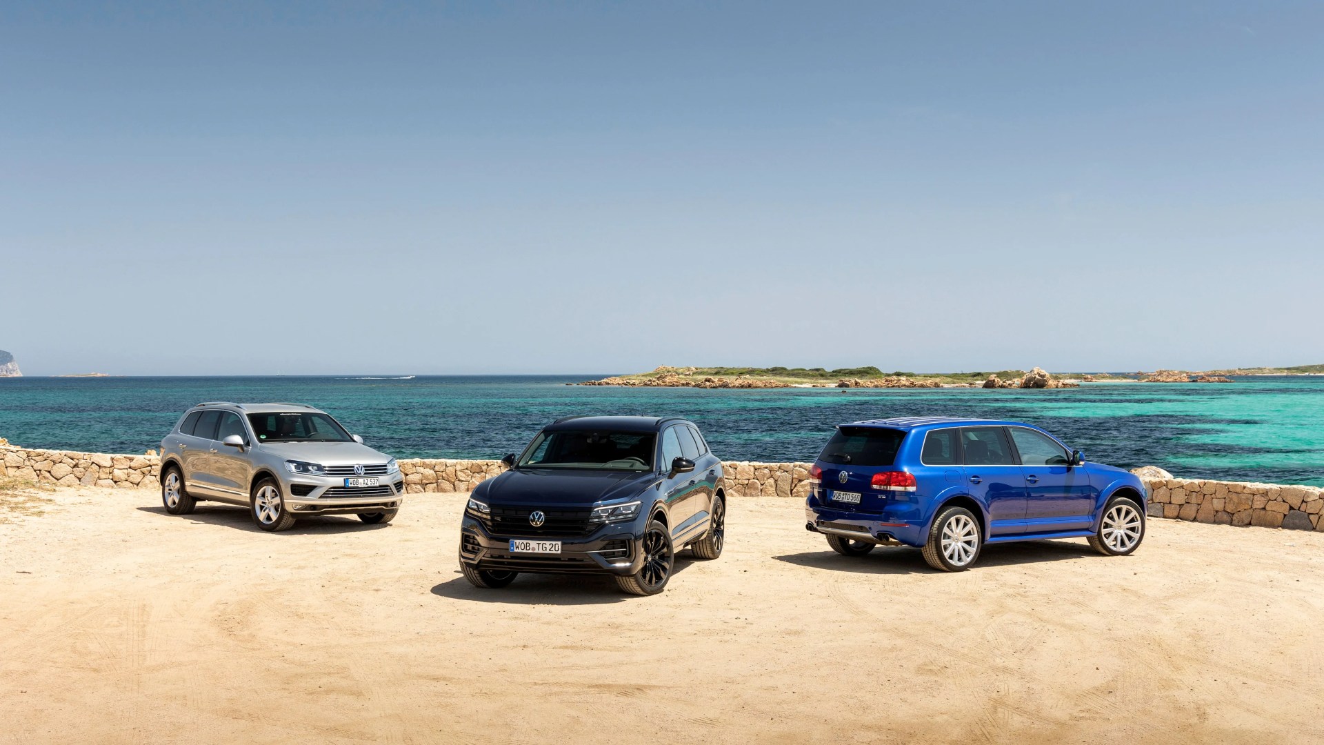 Three Volkswagen SUVs in silver, black, and blue parked on sandy ground near a stone wall with turquoise sea in the background.