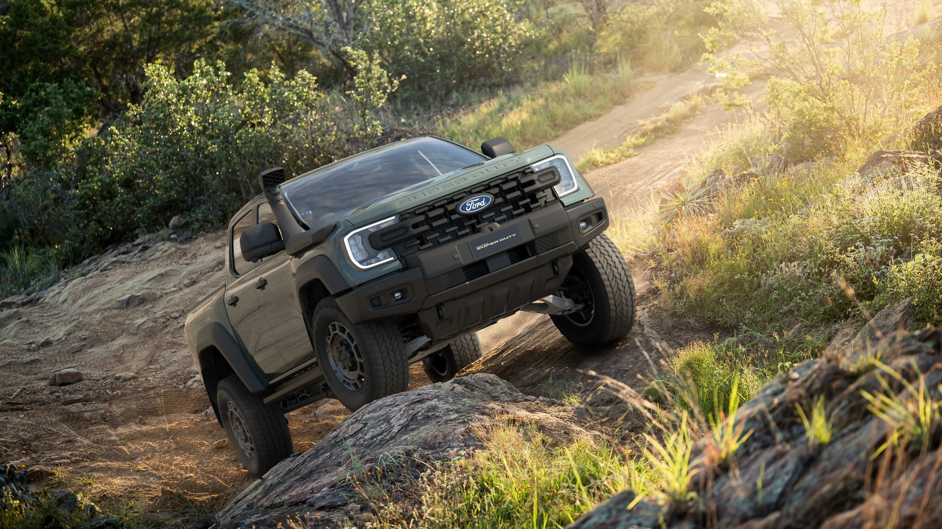Green Ford Super Duty pickup truck climbing rocky off-road trail surrounded by greenery in sunlight.