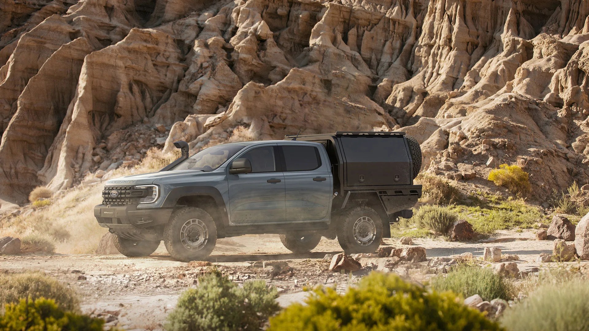 Blue Ford pickup truck with off-road tires and black camper shell parked on rocky desert terrain.