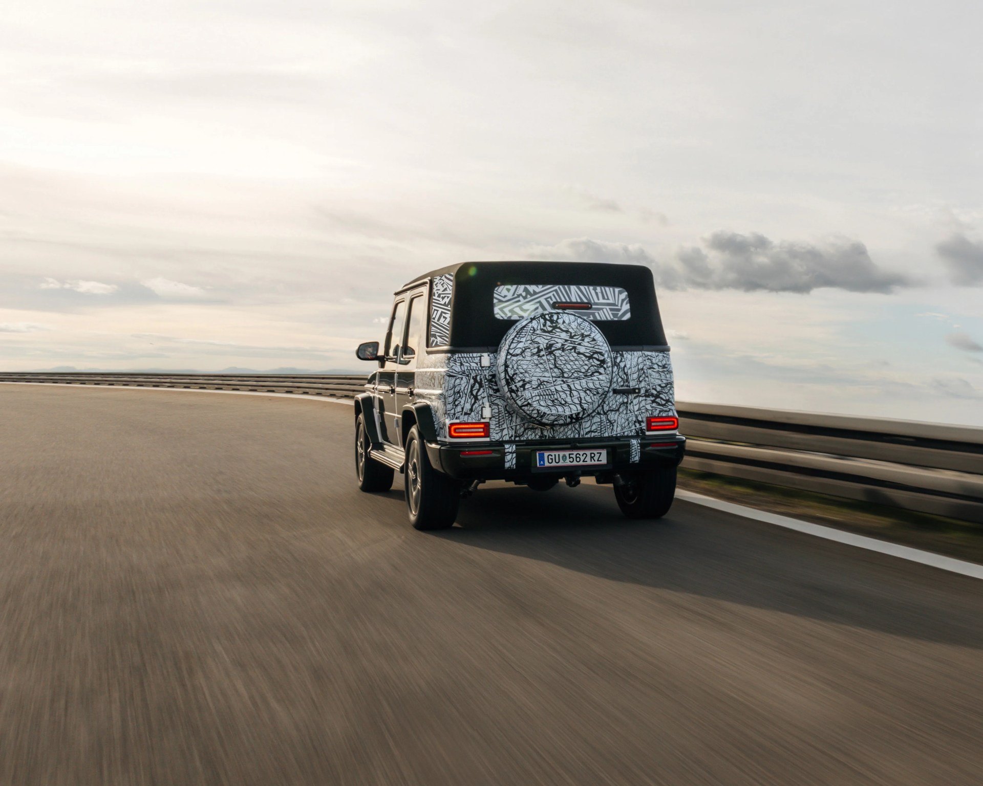 Black and white camouflaged SUV with spare tire cover driving on a curved road under a cloudy sky.