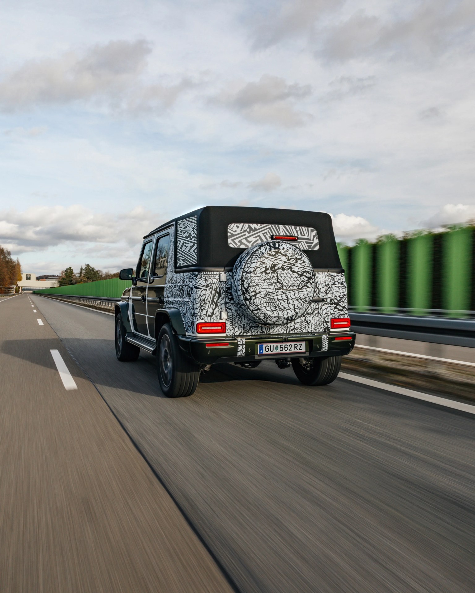 Black and white camouflaged SUV with a soft top driving on a highway with green barriers.