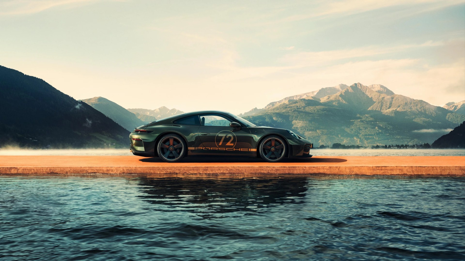 Dark green Porsche 911 with number 72 decal parked on a pier by a lake with mountains in the background.