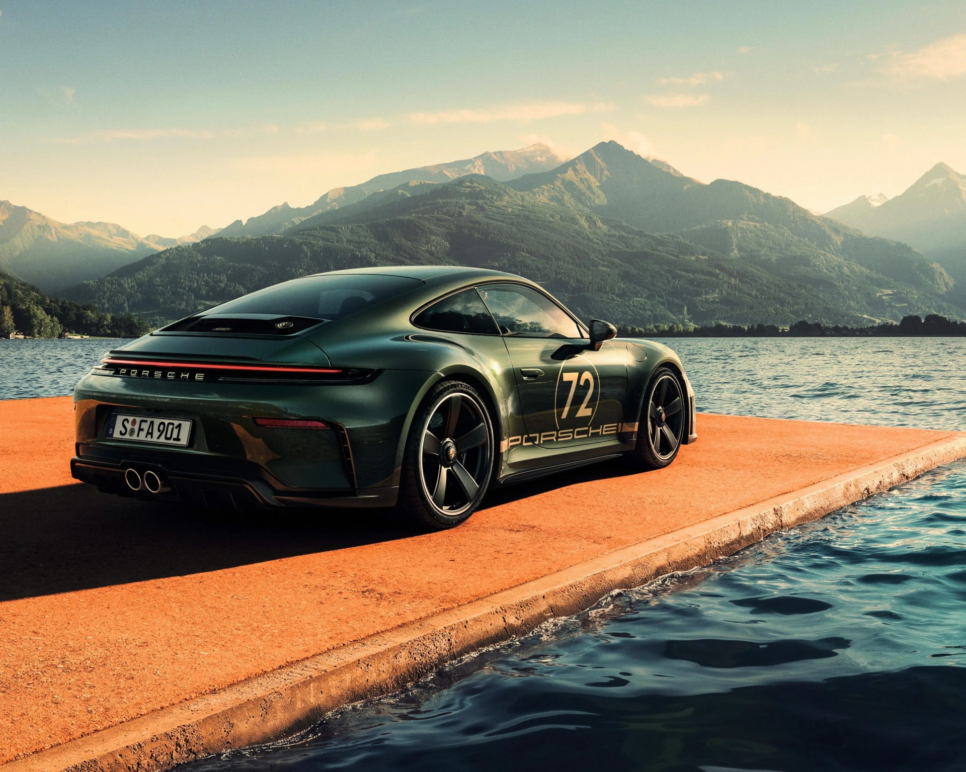 Dark green Porsche 911 with number 72 decal parked on an orange pier by a lake with mountains in the background.