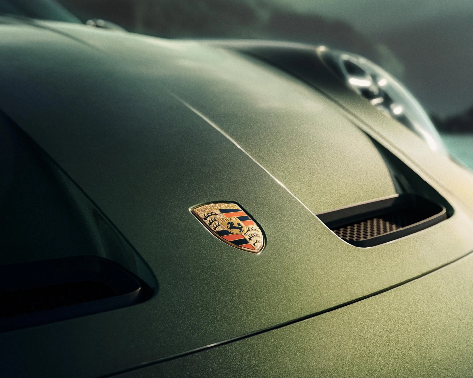 Close-up of a metallic green Porsche car hood with the Porsche emblem and air vents.