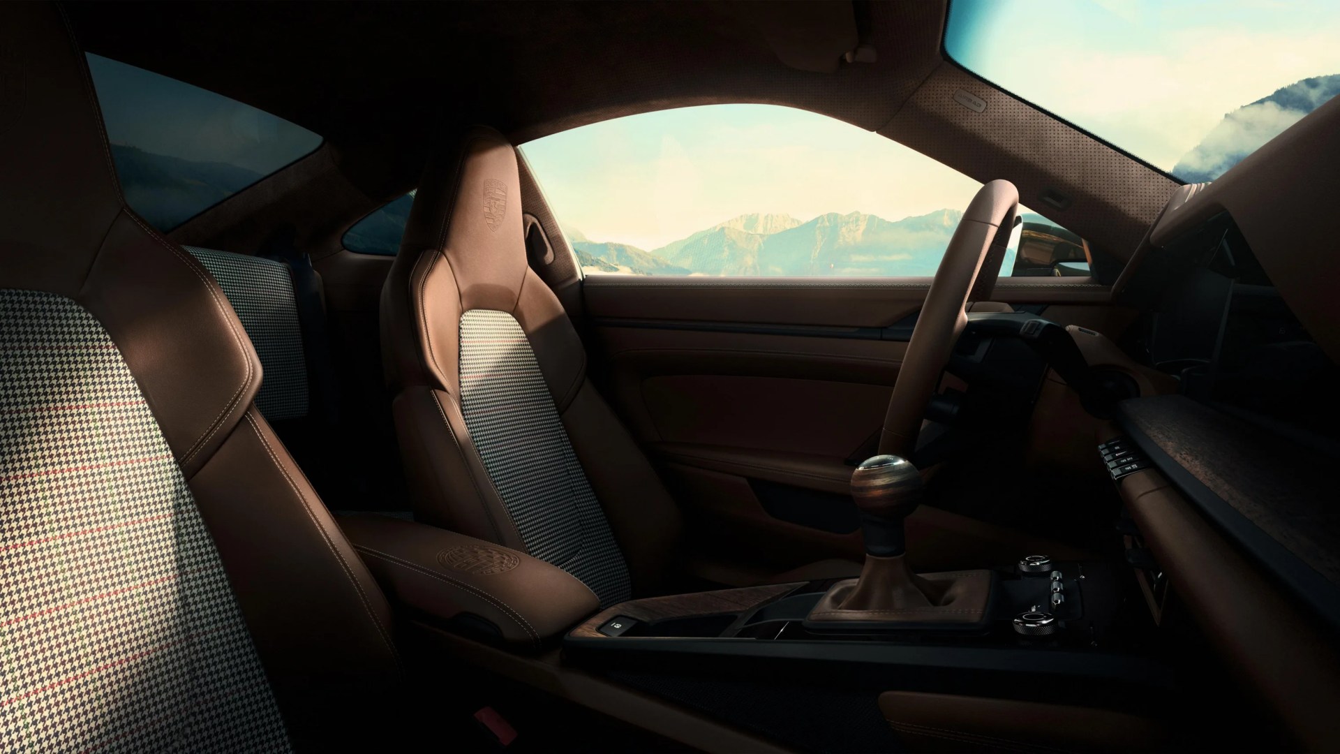 Interior of a car with brown leather and houndstooth fabric seats, a wooden gear shift knob, and a view of mountains through the window.