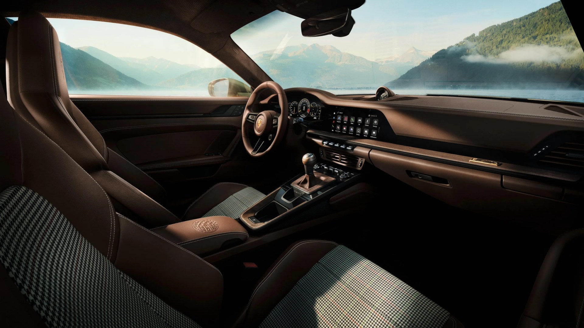 Brown leather and houndstooth fabric car interior with digital dashboard and manual gear shift, overlooking a mountain lake.