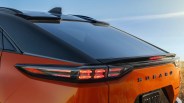 Rear view of an orange Subaru car with illuminated taillights and a black roof spoiler under a clear sky.