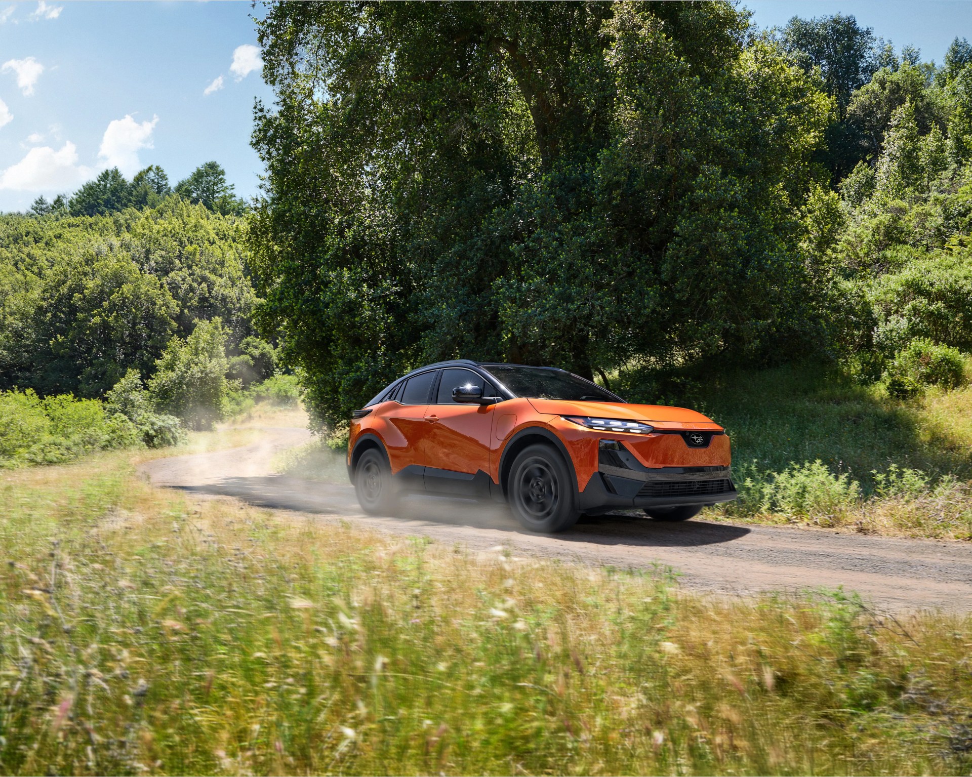 Orange and black electric SUV driving on a dirt road surrounded by green trees and grass.