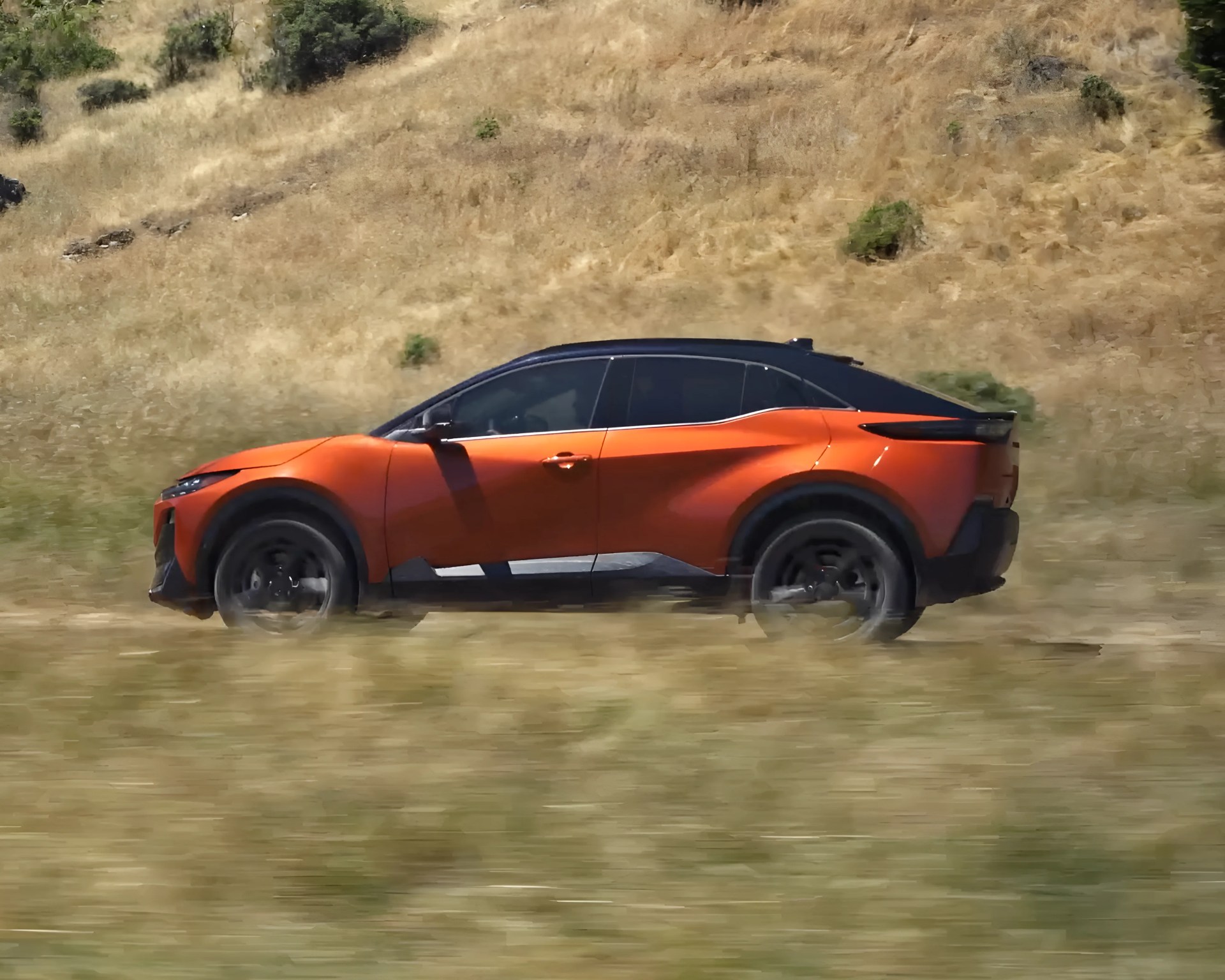 Orange and black electric SUV driving on a grassy dirt road with dry hills in the background.
