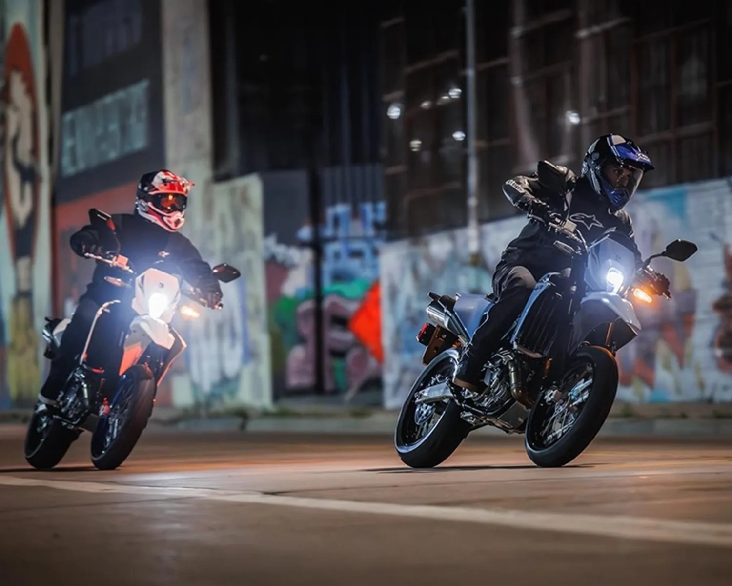 Two motorcyclists wearing helmets and black gear riding on street at night with graffiti-covered walls in background.