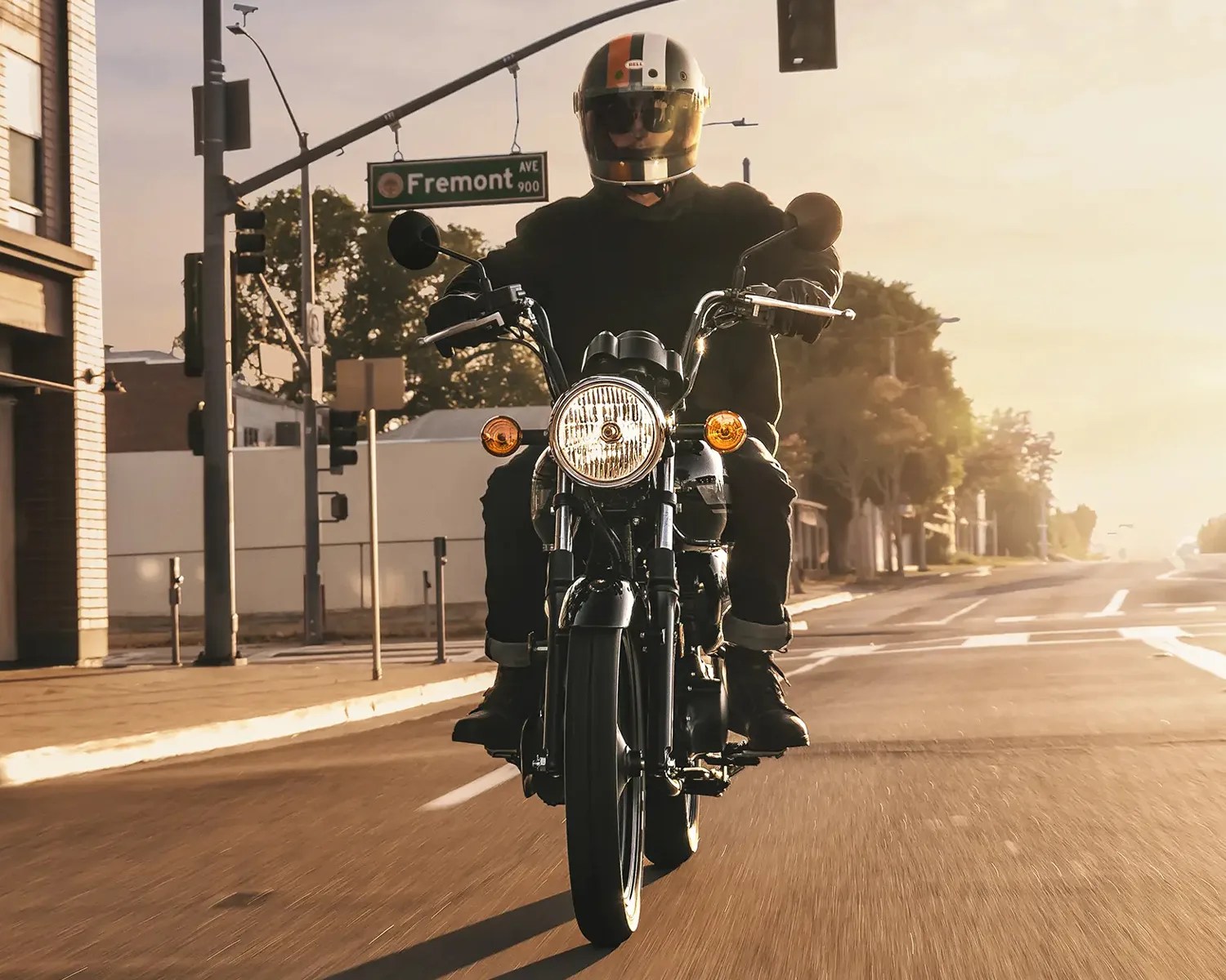 Person wearing a black outfit and helmet riding a black motorcycle on a city street at sunset.
