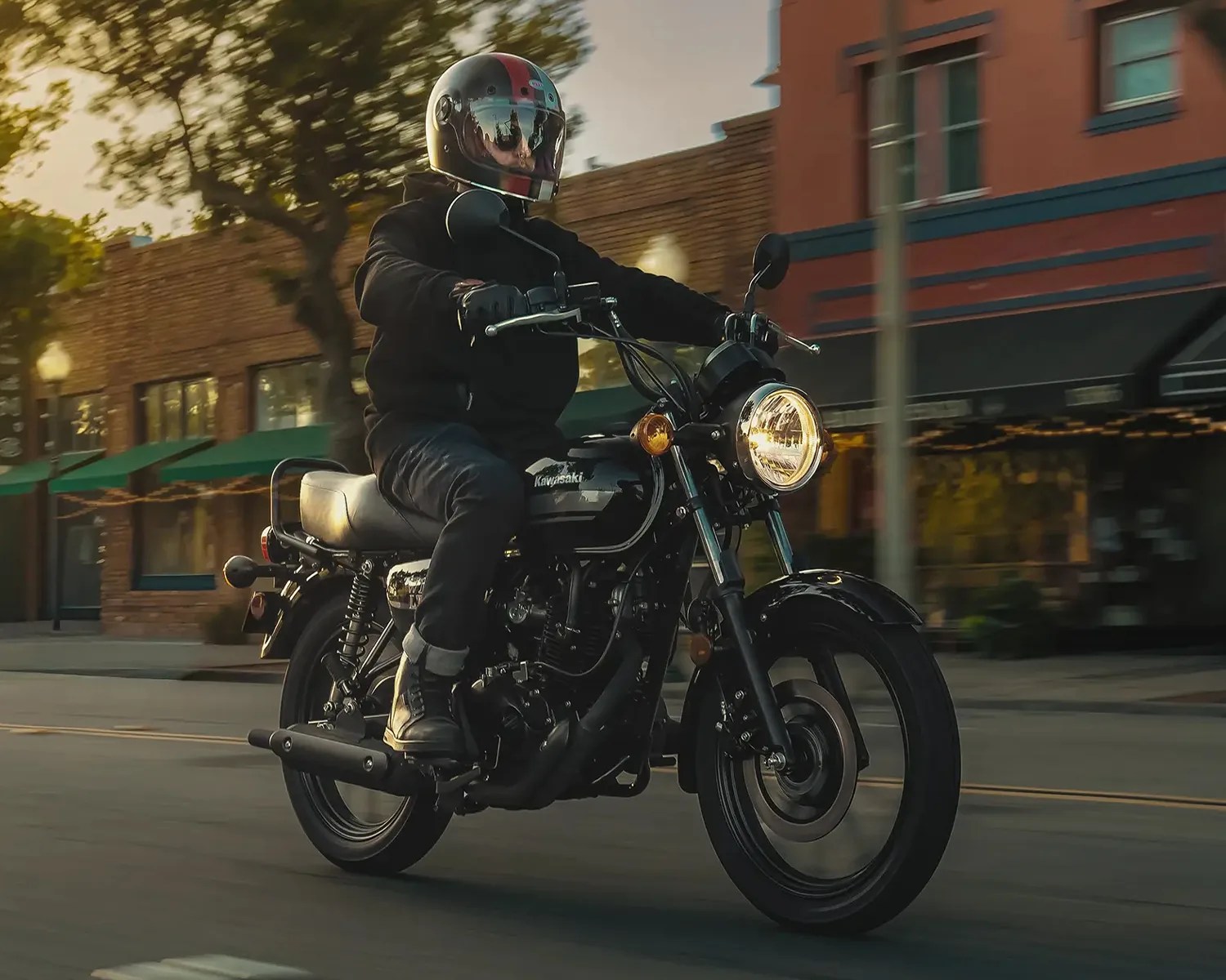 Person wearing black riding gear and helmet riding a black Kawasaki motorcycle on a city street at sunset.