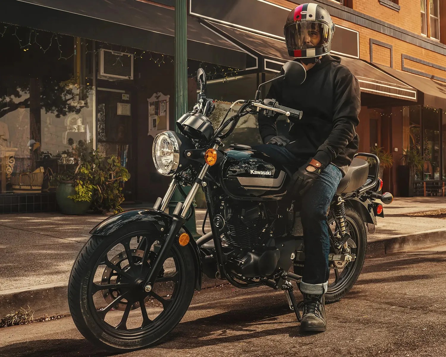 Person wearing a black hoodie and helmet sitting on a black Kawasaki motorcycle on a street.