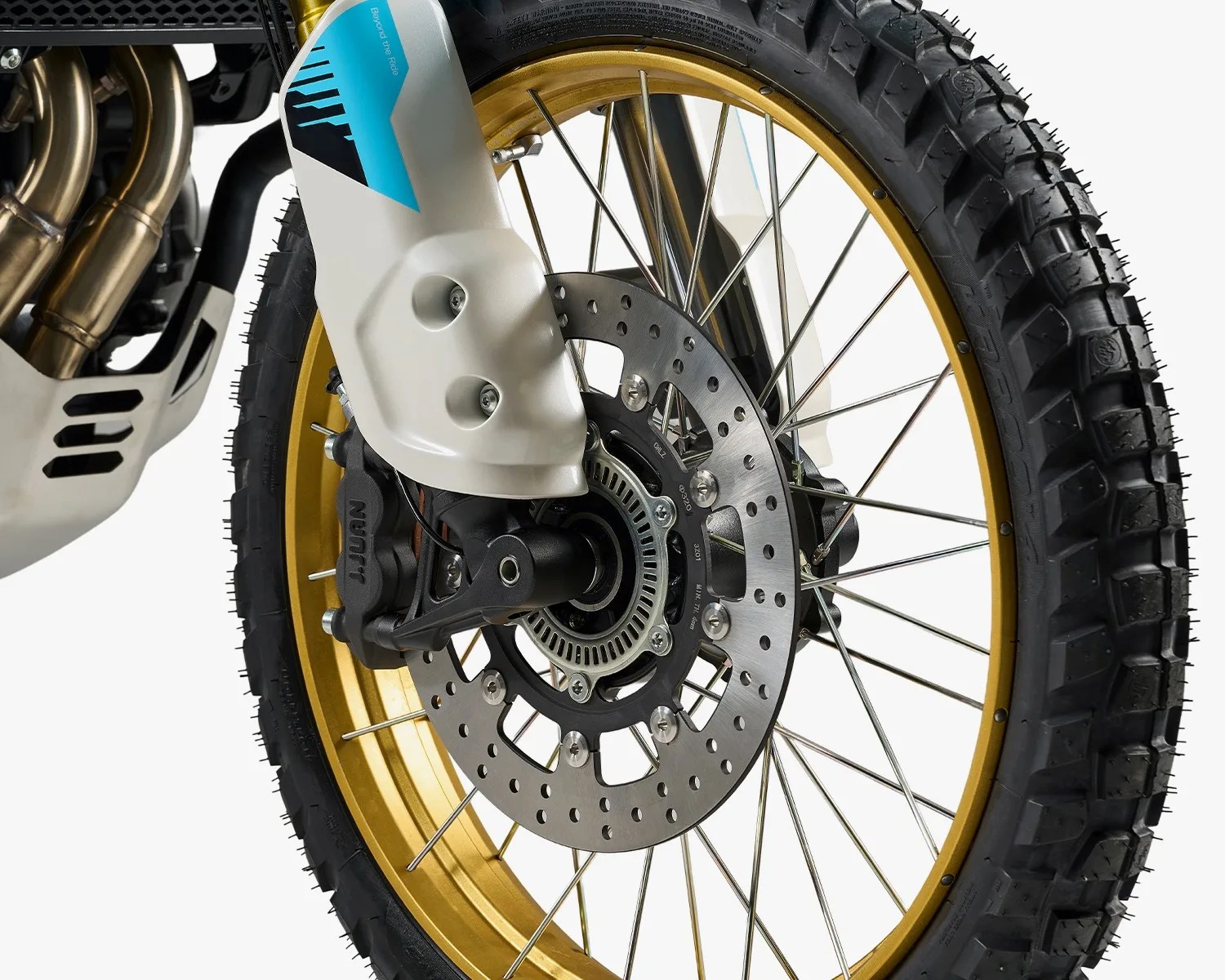Close-up of a motorcycle front wheel with gold rim, knobby tire, disc brake, and white fork guard with blue and black decal.