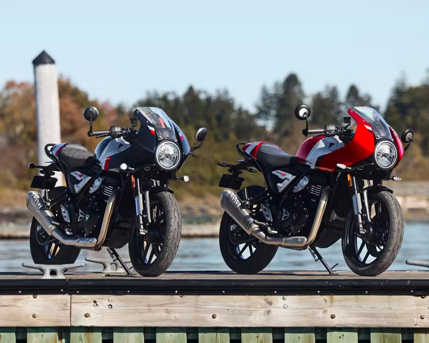 Two retro-style motorcycles, one black and one red, parked side by side on a wooden dock by the water.