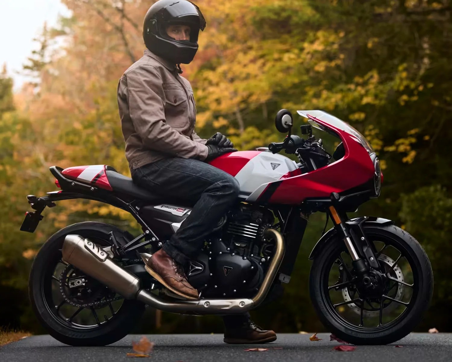 Person wearing a black helmet and brown jacket sitting on a red and white Triumph Thruxton 1200 motorcycle on a road with autumn foliage.