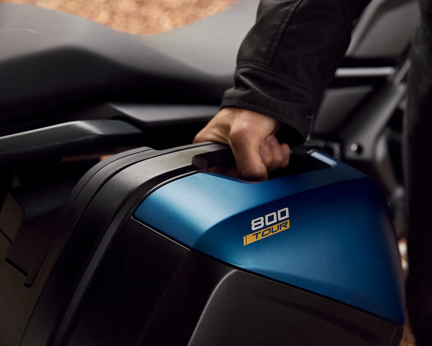 Hand gripping the handle of a blue and black motorcycle side case labeled "800 TOUR.