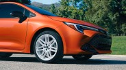Front side view of an orange Toyota car with white alloy wheels parked on a road with trees in the background.