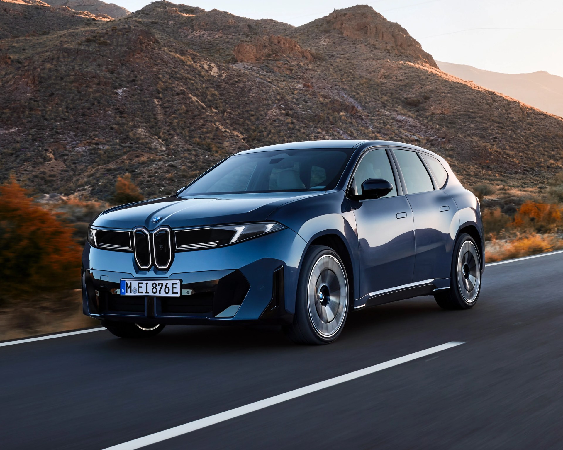 Blue BMW electric SUV driving on a mountain road with rugged hills in the background.