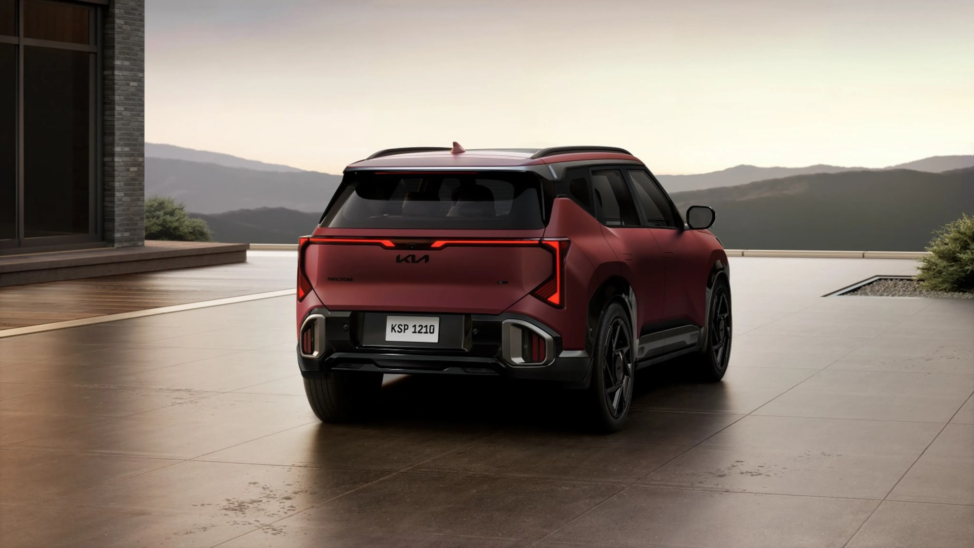 Rear view of a matte red Kia Seltos SUV parked on a tiled patio with mountains in the background.